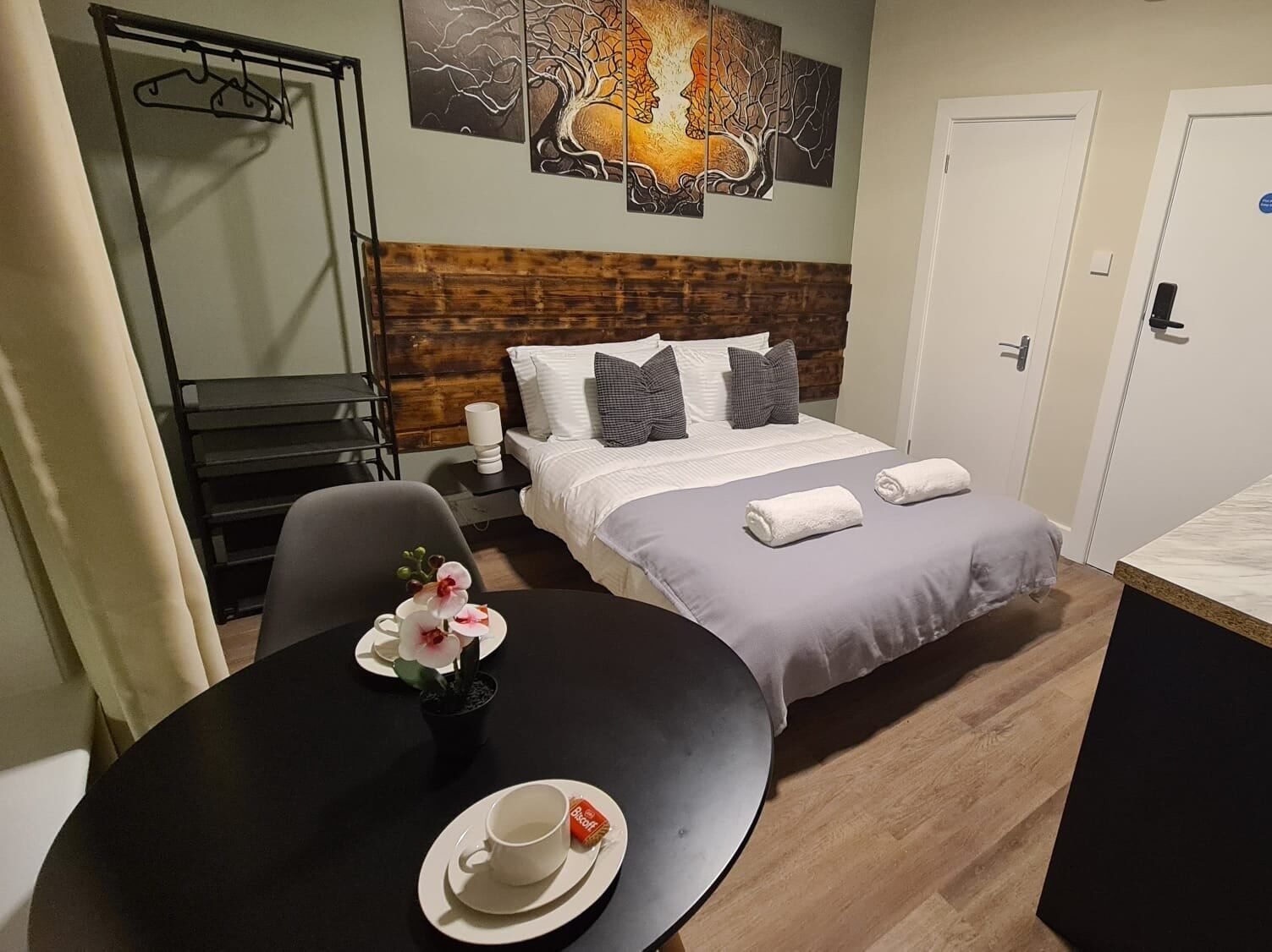 Hotel bed with wooden headboard, white bed linen, gray blanket and folded towels. A table with a flower.